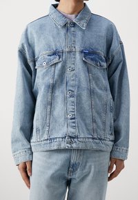 Giacca di jeans azzurro chiaro con due tasche sul petto, chiusura a bottoni e etichetta blu. Caratterizzata da una vestibilità comoda e un texture leggermente vissuta.