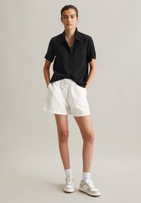 Schwarzes Kurzarm-Hemd mit Knopfverschluss kombiniert mit weißen Shorts mit Kordelzug und weißen Sneakers. Das Modell steht mit den Händen in den Taschen vor einem neutralen Hintergrund.
