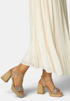 Piernas con falda plisada color crema, usando sandalias beige de tacón grueso con tira al tobillo, plataforma texturizada tejida y tira superior con joyas multicolores.