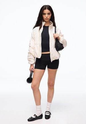 Ung kvinde i sort crop top og shorts med beige jakke, holder sorte høretelefoner og en lille sort skuldertaske, står på hvid baggrund.