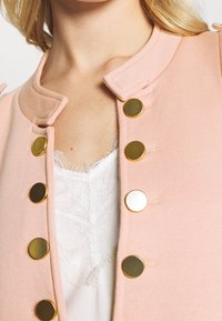 Veste rose clair à col mandarin, ornée de boutons ronds dorés et argentés, portée sur un haut en dentelle blanche aux bords effilochés.