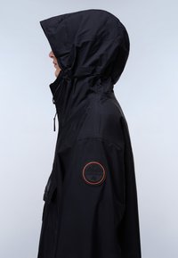 Άτομο που φοράει μαύρο μπουφάν με κουκούλα και ένα στρογγυλό ένθετο πορτοκαλί περιγράμματος στο πάνω μέρος του μπράτσου, στρέφεται αριστερά μπροστά σε απλό φόντο.