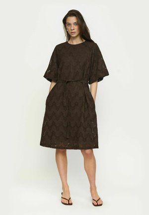 Naine seisab otse, kandes põlveni ulatuvat tumepruunist pitskleiti lühikeste varrukatega, vööga talje ümber ja sandaale, heleda ühevärvilise tausta ees.