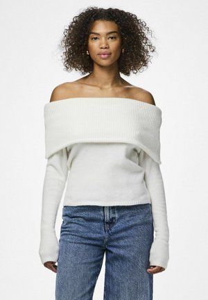 Maglione bianco off-the-shoulder con colletto a coste e maniche lunghe, abbinato a jeans azzurri rilassati. Texture morbida, vestibilità casual.