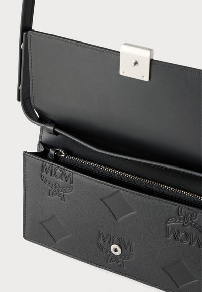 Μαύρη δερμάτινη τσάντα MCM για κράτημα στον ώμο με ανάγλυφα λογότυπα, ασημένια κούμπωμα και εσωτερικό διαμέρισμα με φερμουάρ.
