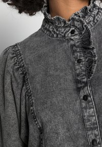 Närbild av en bleknad svart denimjacka med volangkrage och svarta tryckknappar på en person med lockigt hår.