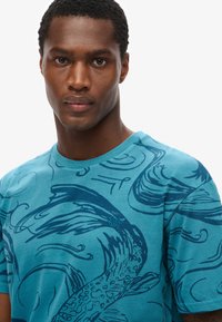 Camiseta turquesa con patrones de peces y olas en azul oscuro. Diseño de cuello redondo, mangas cortas, hecha de tela de algodón suave.