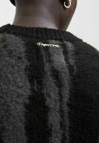 Nahaufnahme des Rückens einer Person, die einen schwarzen, flauschigen Pullover mit dem Logo "Project One" nahe dem Kragen trägt, sowie einem sichtbaren silbernen Ringohrring.