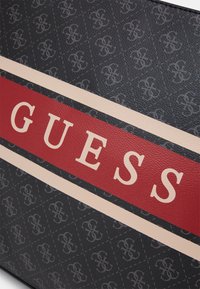 Czarny teksturowany materiał z powtarzającym się wzorem logo, ozdobiony wyrazistym czerwonym paskiem z napisem "GUESS" w kolorze kremowym.