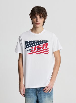 Młody mężczyzna o potarganych włosach nosi białą koszulkę z stylizowaną flagą Ameryki i napisem "Made in USA" w kolorze czerwonym i niebieskim.