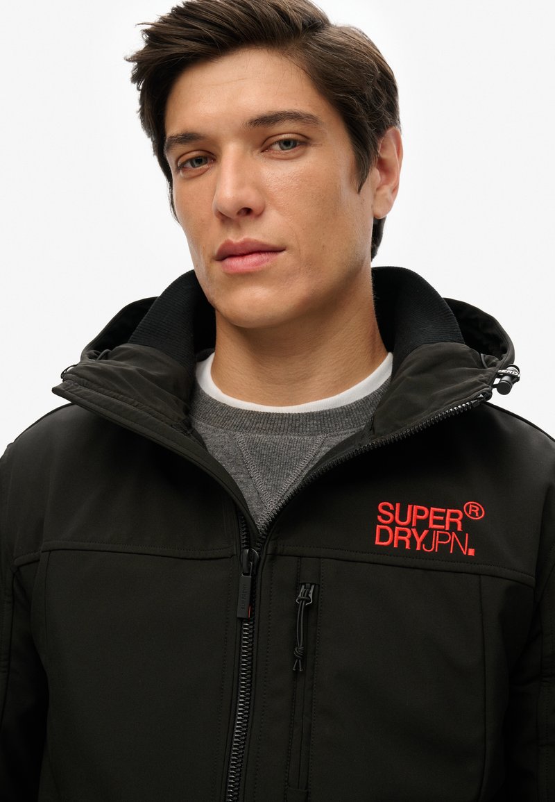 Zwarte jas met een hoge kraag, voorrits en geborduurd rood "SUPER DRY JPN" logo. Grijze sweatshirt zichtbaar eronder. Vriendelijke textuur.