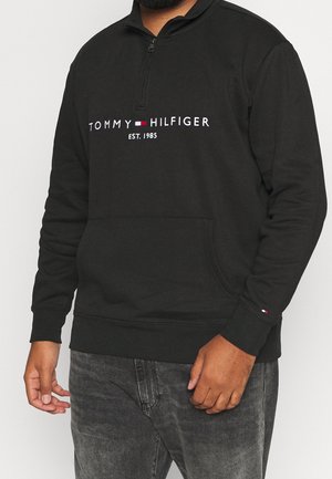 Mann trägt einen schwarzen Tommy Hilfiger Pullover mit einem Viertel-Reißverschluss, Vordertasche und Logotext auf der Brust, kombiniert mit ausgewaschenen schwarzen Jeans.