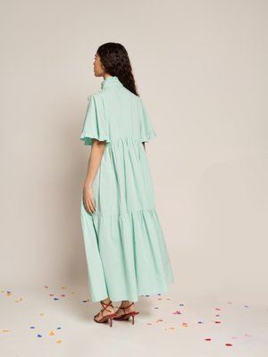 Rochie maxi verde mentă, cu volane, cu mâneci scurte, guler înalt și fustă în straturi. Purtați cu sandale roșii cu sireturi pe un podea presărată cu confetti.