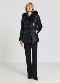 Giacca imbottita nera con colletto in ecopelliccia, vita con cintura e tasche con cerniera. Indossata sopra pantaloni neri a gamba larga.