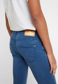 Person trägt eine blaue, eng anliegende Jeans mit Gesäßtaschen und einem tanfarbenen Leder-Label, kombiniert mit einem hellgrauen, in die Hose gesteckten Shirt.