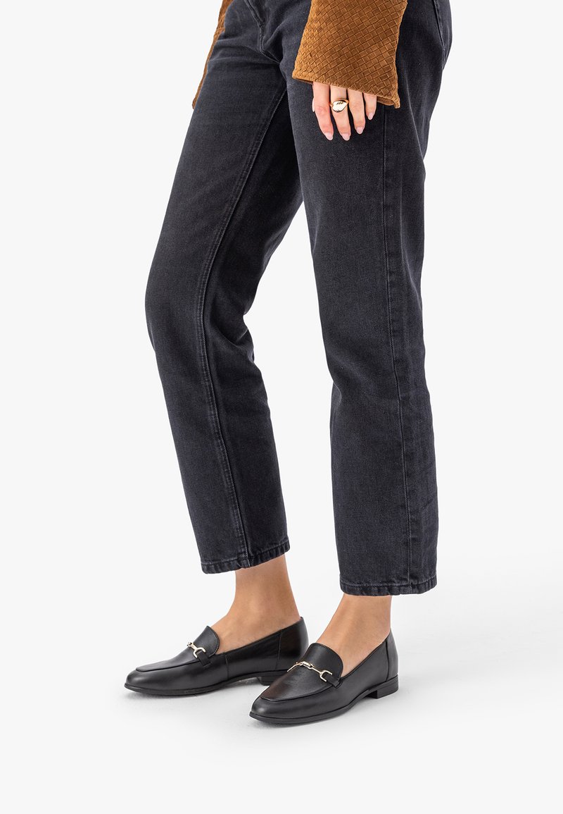 Černé kožené mokasíny se zlatými přezkami, nošené s krátkými černými džínami a hnědým prošívaným rukávem viditelným na bílém pozadí.