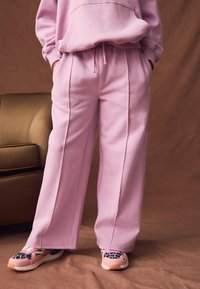 Sudadera con capucha de forro polar rosa y pantalones a juego con costuras laterales. Las zapatillas presentan detalles de estampado animal y una base rosa vibrante. Sofá marrón en el fondo.