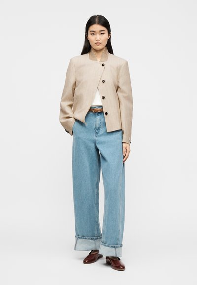 Kvinde iført beige knappet jakke, lyseblå vide bukser med opbuk, brunt bælte og brune sko, stående mod en hvid baggrund.