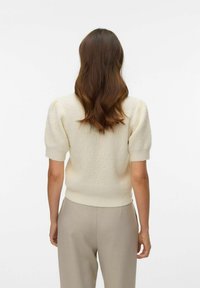 Pull en tricot texturé de couleur crème avec manches courtes bouffantes et taille cintrée ; porté avec un pantalon beige clair.