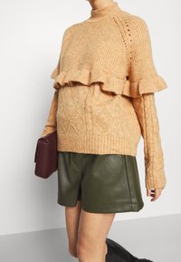 Strickpullover in warmem Beige, mit gerafftem Saum, Zopfmuster und hohem Kragen, kombiniert mit dunkelgrünen Lederhosen und einer burgunderfarbenen Tasche.