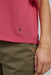 Kratký rukáv, červený top s texturovaným povrchem; na spodním lemu má malé metalické logo. Spárováno s olivově zelenými kalhotami a zlatým náramkem.