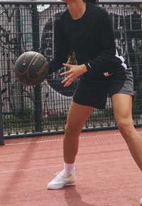 Camisa negra de manga larga, pantalones cortos deportivos negros y grises, zapatillas blancas, sosteniendo un balón de baloncesto marrón en una cancha roja con un fondo de malla.
