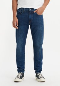 Moški v temno modrih džinsih in črnih visokih supergah stoji z eno roko v žepu ob belem ozadju.