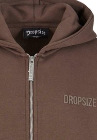 Hnedá mikina s kovovým zipsom, vyrobená z mäkkej tkaniny, s textom "DROPSIZE" v kontrastnej farbe a veľkou kapucňou.