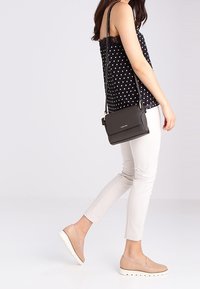 Borsa a tracolla nera con texture liscia, forma rettangolare e hardware argentato. Indossata con un top nero a pois e pantaloni bianchi.