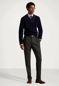 Homem com óculos, suéter azul marinho em malha de corda com decote em V sobre uma camisa e gravata azuis, calças cinzas, cinto e sapatos castanhos, de pé com uma mão no bolso.