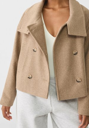 Veste en laine beige à double boutonnage portée sur un top blanc et un pantalon gris clair, montrant le torse et les bras sur un fond uni.