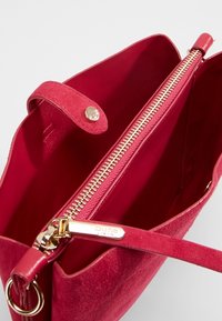 Punainen samettinen käsilaukku, jossa on teksturoitu pinta, kultainen vetoketju, metalliosat ja nappikatkaisu. Sisäpuoli vuorattu vastaavalla punaisella.