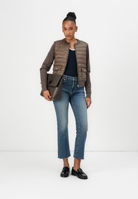 Braune gepolsterte Bomberjacke über einem marineblauen Hemd, kombiniert mit hellblauen, cropped Jeans und schwarzen Slippern. Das Model hält eine schwarze Handtasche.