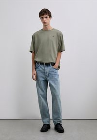 Camiseta de algodón verde oliva, corte holgado, con logo en el pecho. Vaqueros azul claro de pierna recta, lavado desgastado, cinturón oscuro y zapatos de cuero negros.