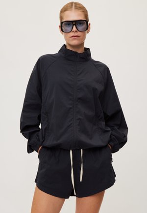 Mujer con chaqueta negra grande con cremallera, shorts a juego con cordón blanco y grandes gafas de sol negras, de pie con las manos en los bolsillos.