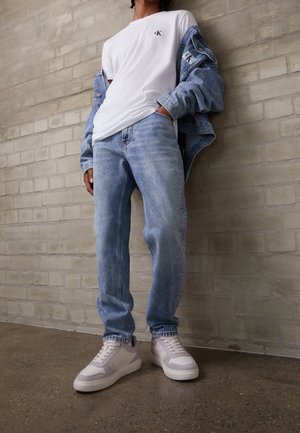 Personne portant un jean bleu clair, des baskets blanches, un t-shirt blanc Calvin Klein et une veste en jean suspendue sur les épaules devant un mur en briques.