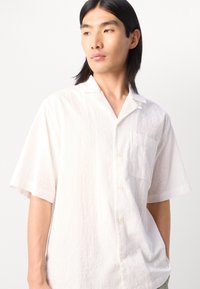Short-sleeve white shirt in textured fabric with a subtle wave pattern, featuring a collar and a left chest pocket, button-front design.