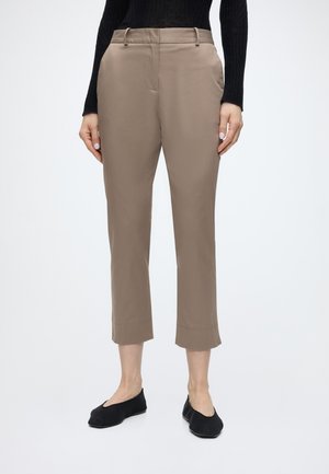 Personne portant un pantalon beige raccourci, un haut noir à manches longues et des ballerines noires, montrée des épaules aux pieds sur un fond uni.