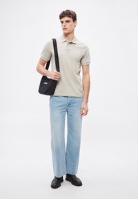 Ανοιχτό μπεζ πόλο πουκάμισο, ανοιχτό μπλε τζιν παντελόνι φαρδύ στο πόδι, μαύρα μποτάκια αστραγάλου και μια μικρή μαύρη τσάντα χιαστί με λευκό λογότυπο, στέκεται μπροστά σε λευκό φόντο.