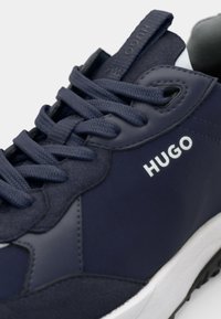 Mornarsko modri športni čevlji z teksturirano usnjeno in blago površino. Imajo bel logo, okrogle vezalke in belo srednjo podplato. Vidna gumijasta podplata.