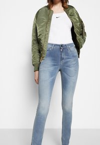 Donna che indossa jeans skinny a vita alta blu chiaro, canotta bianca Nike e giacca bomber verde oliva all'interno contro uno sfondo bianco.