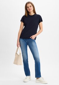 Marineblauwe t-shirt met korte mouwen, gecombineerd met lichtblauwe spijkerbroek en witte sneakers. Het model houdt een beige handtas vast met een gestructureerd ontwerp.