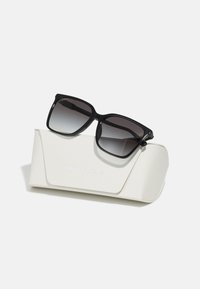 Black sunglasses with gradient lenses resting on a white textured case featuring embossed branding. Square frame design.