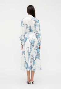 Λευκό φόρεμα πουκάμισο με μπλε λουλουδάτο σχέδιο, μακριά μανίκια, σφηκοφωλιά στη μέση και αέρινη φούστα, φτιαγμένο από ελαφρύ ύφασμα. Ορατές γόβες.