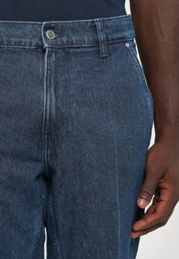 Jeans de mezclilla azul oscuro con cierre de botón de metal, parte de una blusa negra, y una mano de piel oscura descansando junto al bolsillo.