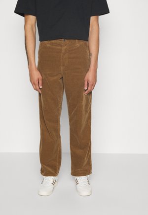 Persona con pantalones de pana marrones, camiseta negra y zapatillas blancas de pie contra un fondo liso.