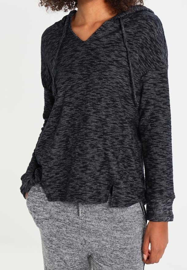 Femme portant un sweat à capuche chiné gris foncé avec cordons et un pantalon de jogging chiné gris clair, mains dans les poches.