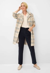 Ruiten wollen jas in beige en grijstinten, met een notch kraag, sluiting met één knoop, gedragen over een lichte blouse en donkere broek.