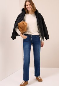 Donna che sorride, indossando jeans blu scuro, un maglione beige e una giacca nera appoggiata sulle spalle, con una borsa marrone in mano, in piedi su uno sfondo chiaro.