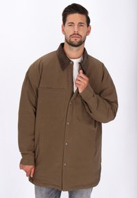 Mann mit Bart, der eine braune, geknöpfte Jacke mit dunklem Kragen und hellhem Hemd trägt, die Jacke anpasst, neutraler Hintergrund.